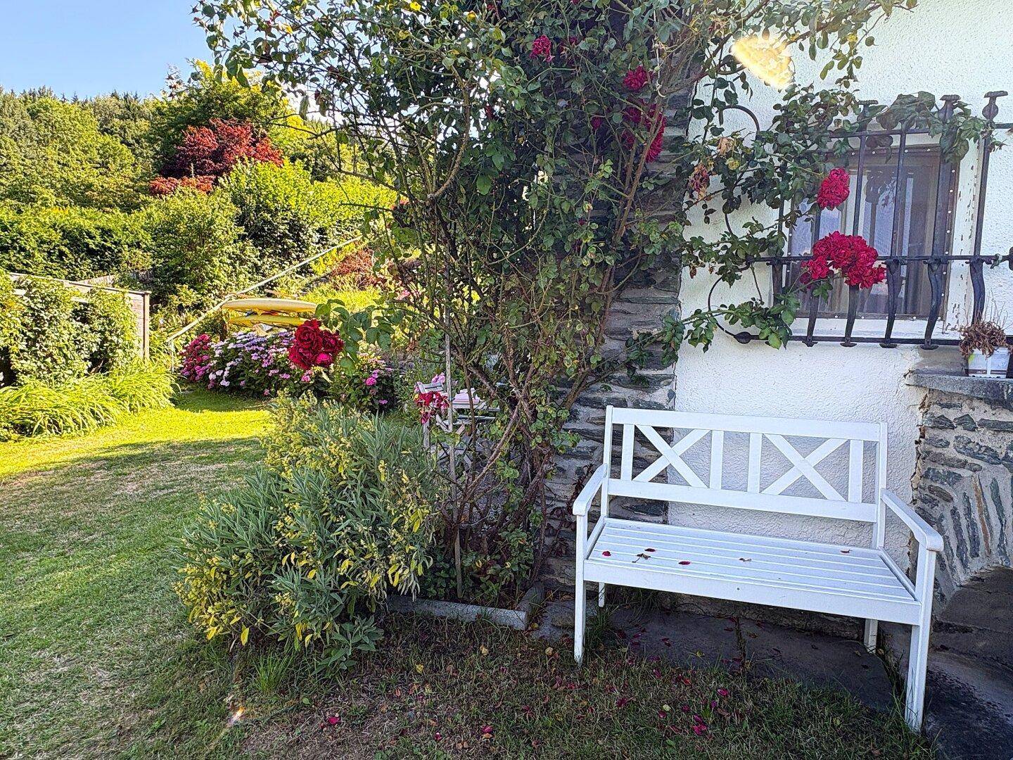 Idyllischer Gartenbereich mit weißer Holzbank, blühenden Pflanzen und üppigem Grün am Haus.