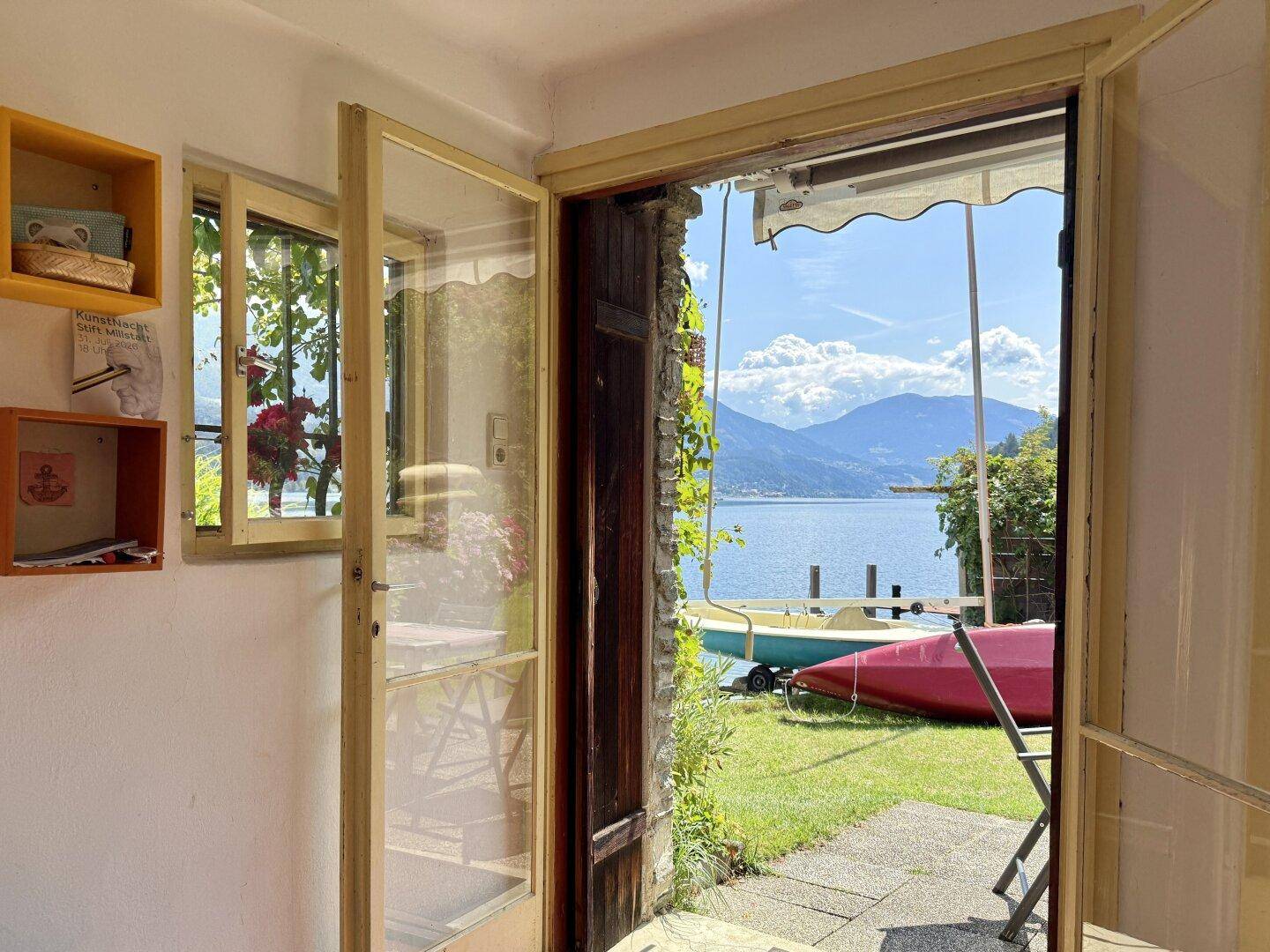 Blick aus dem Innenraum durch die offene Tür auf den See, ein Boot und die umliegenden Berge.