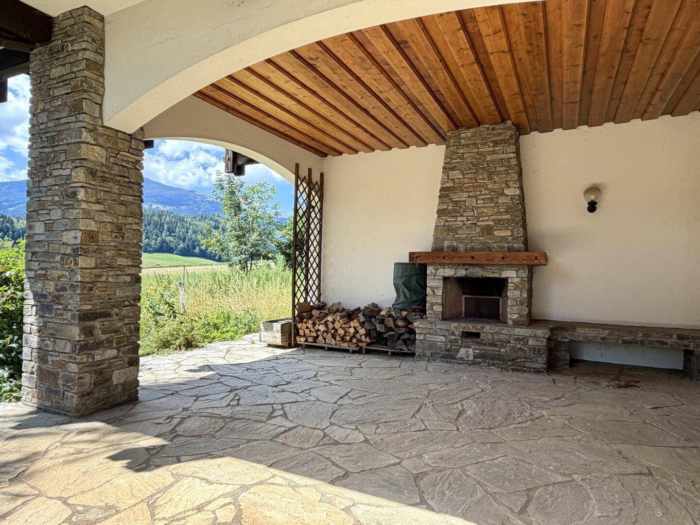 Überdachte Steinterrasse mit Außenkamin, Holzstapel und Bergblick.