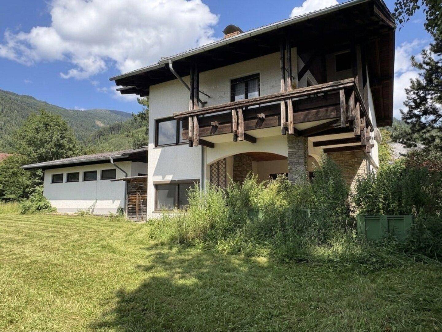 Traditionelles Haus mit Balkon, umgeben von üppiger Vegetation und Bergen.