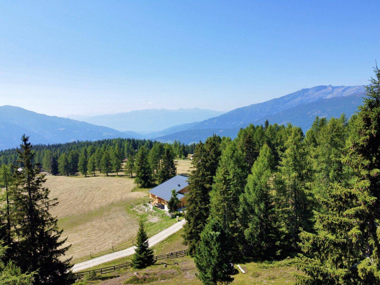 Panoramablick über ein Tal mit einem Haus, umgeben von Wäldern und Wiesen, unter blauem Himmel.