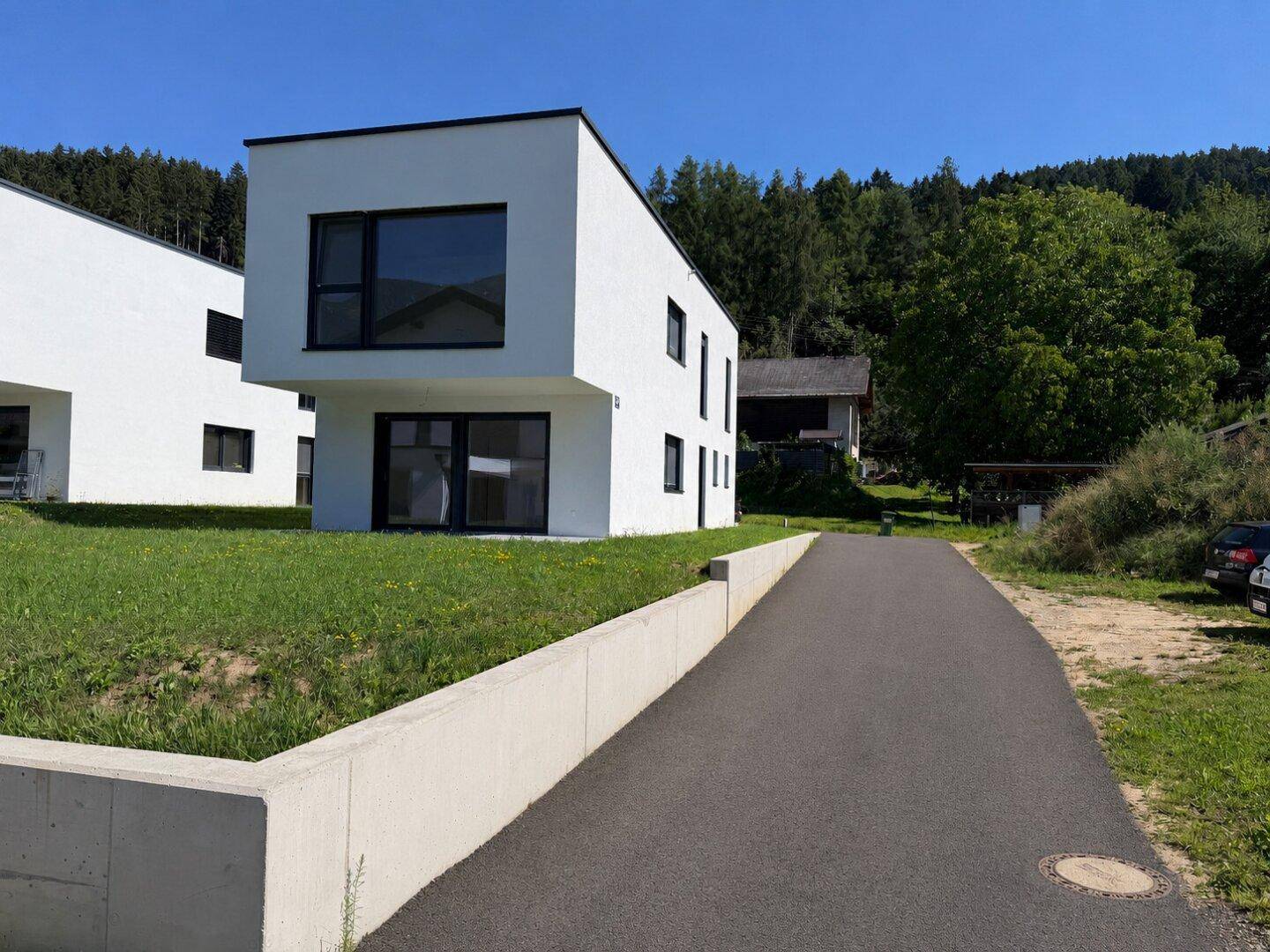 Modernes Einfamilienhaus mit weißer Fassade und Zufahrt, umgeben von grüner Natur.