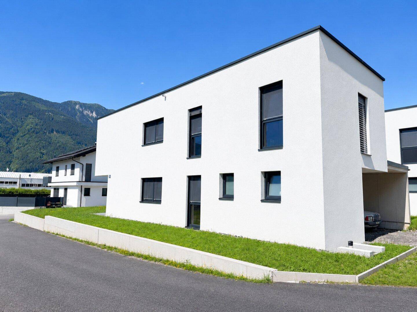 Modernes Wohnhaus mit weißer Fassade und dunklen Fensterrahmen, eingebettet in eine grüne Landschaft.