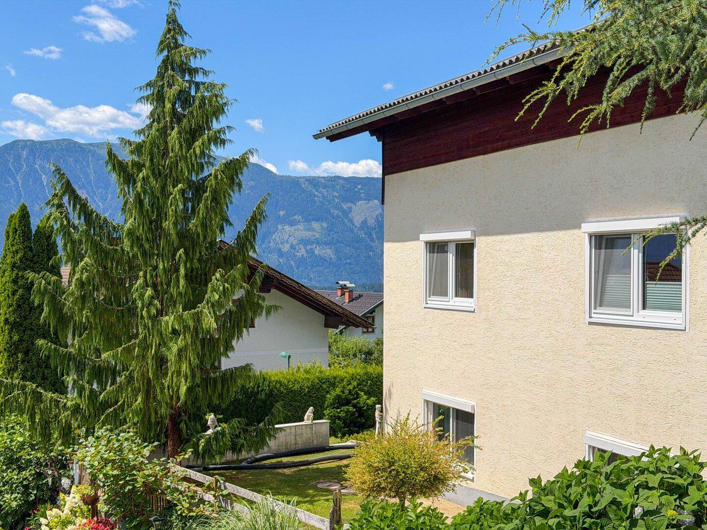 Seitenansicht des Hauses mit Rollläden, umgeben von Grün und Bergblick.