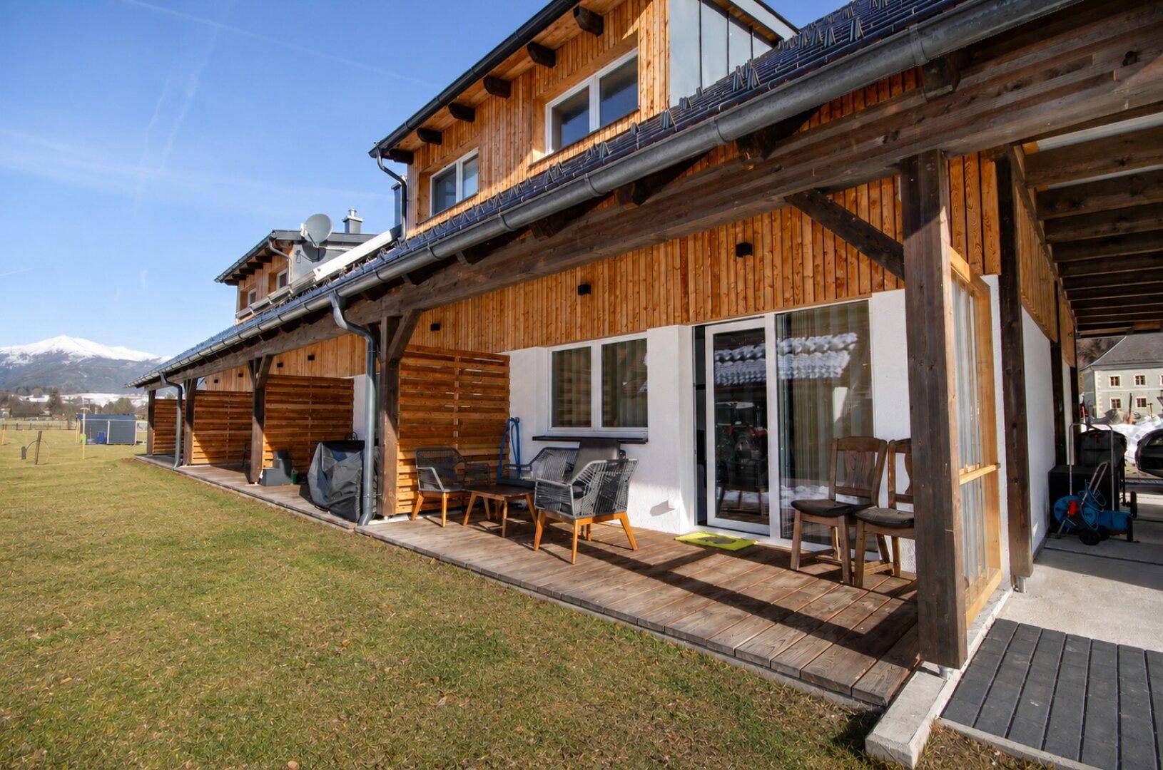 Überdachte Terrasse mit Holzboden und Sitzgelegenheiten, die zu einem grünen Rasen und Bergblick führt.