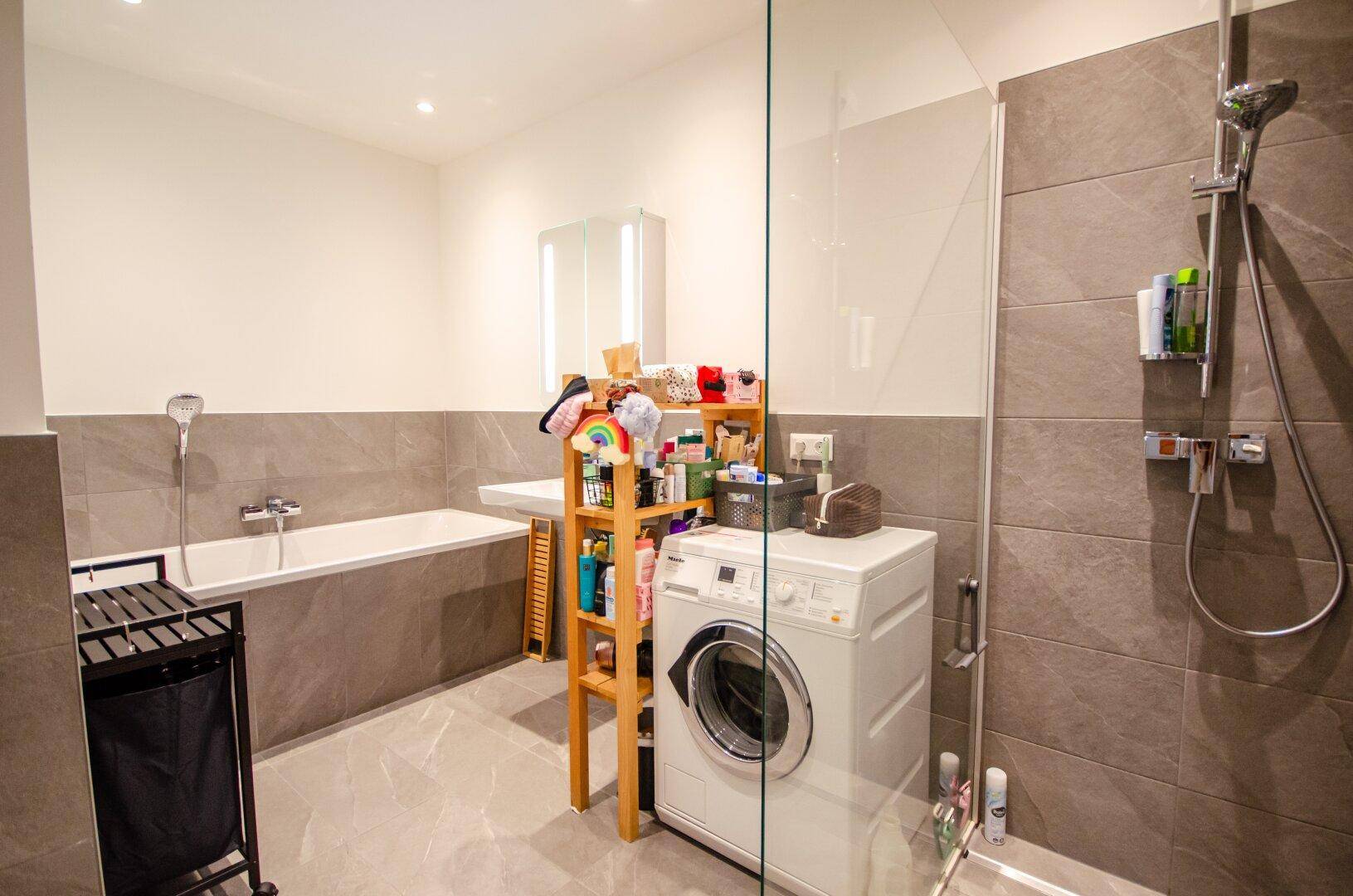 Modernes Badezimmer mit Badewanne, begehbarer Dusche, Waschmaschine und grauen Fliesen.