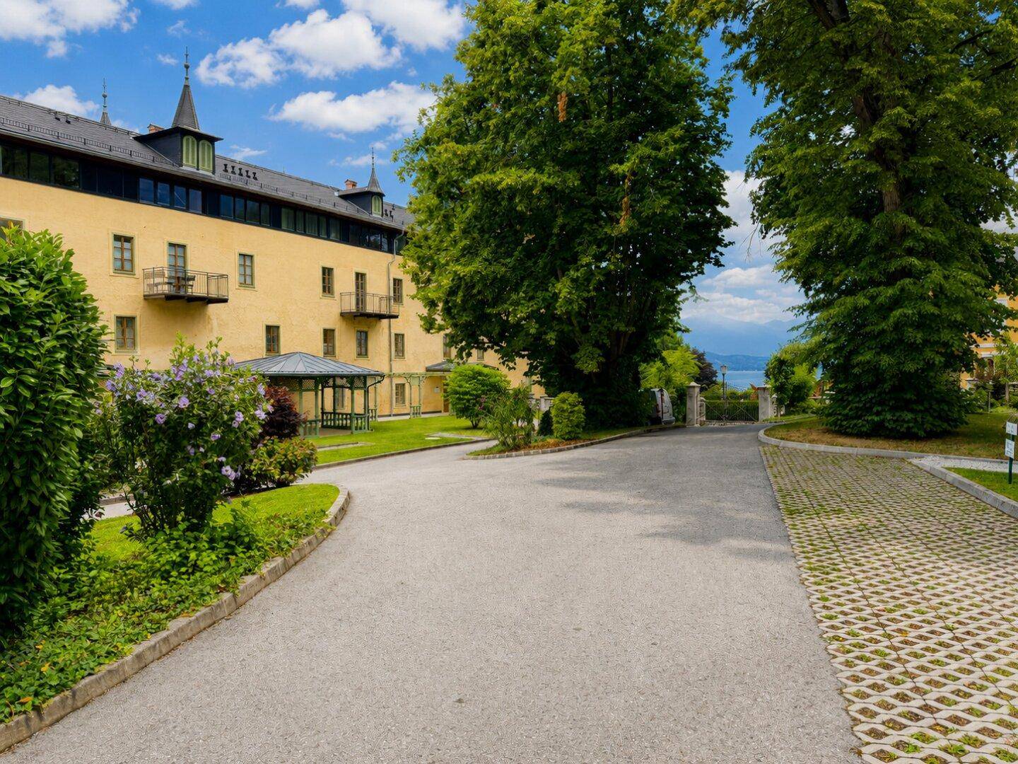Weitläufige Außenanlage des Gebäudes mit großen Bäumen, Kiesweg und einem entfernten Blick auf den See.