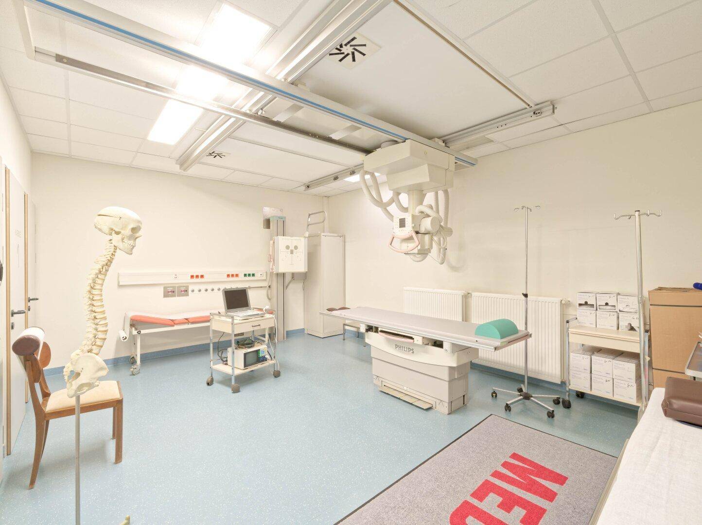 Medizinischer Untersuchungsraum mit Röntgengerät, Untersuchungsliege und einem Skelettmodell.