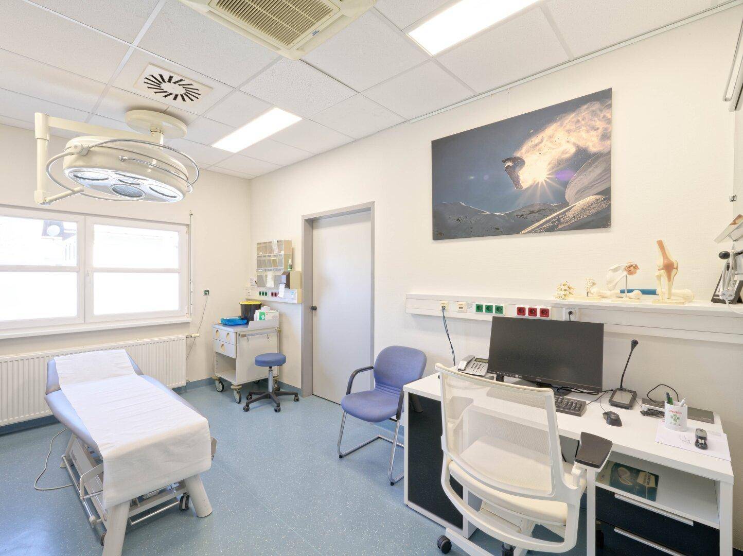Heller Untersuchungsraum mit Liege, Schreibtisch, Computer und medizinischen Instrumenten.