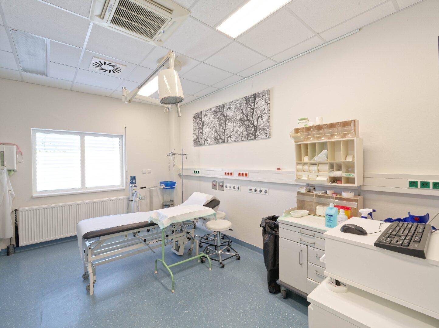 Medizinischer Behandlungsraum mit Untersuchungsliege, Schränken und einem großen Fenster.