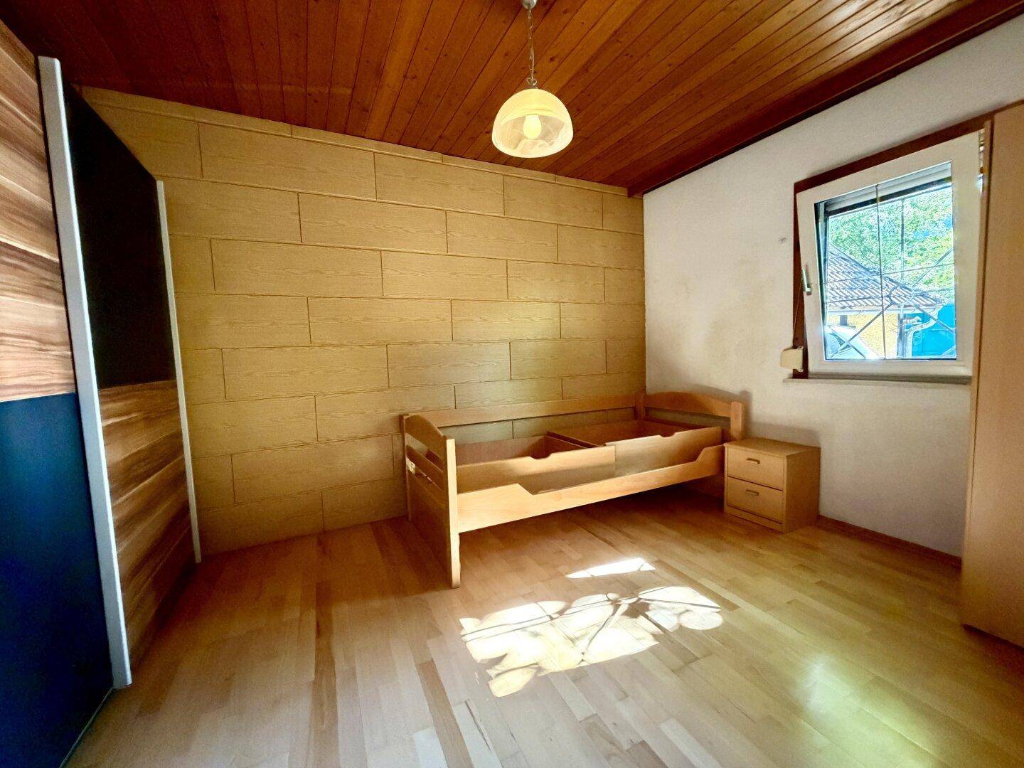 Schlafzimmer mit Holzboden, Holzdecke und einer Holzwandverkleidung, hell durch das Fenster.