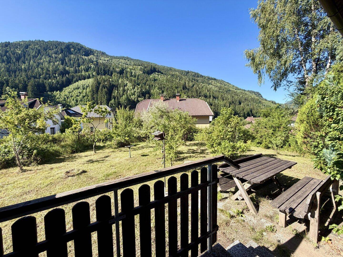Rustikaler Picknickbereich im Garten mit Holztisch und Bänken, umgeben von Natur und Bergblick.