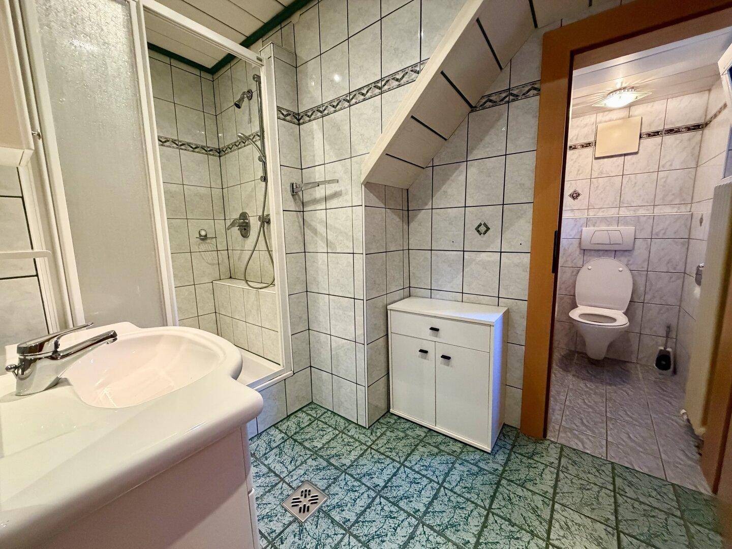 Gefliestes Badezimmer mit Dusche, Waschbecken und Dachschräge, angrenzend an eine separate Toilette.