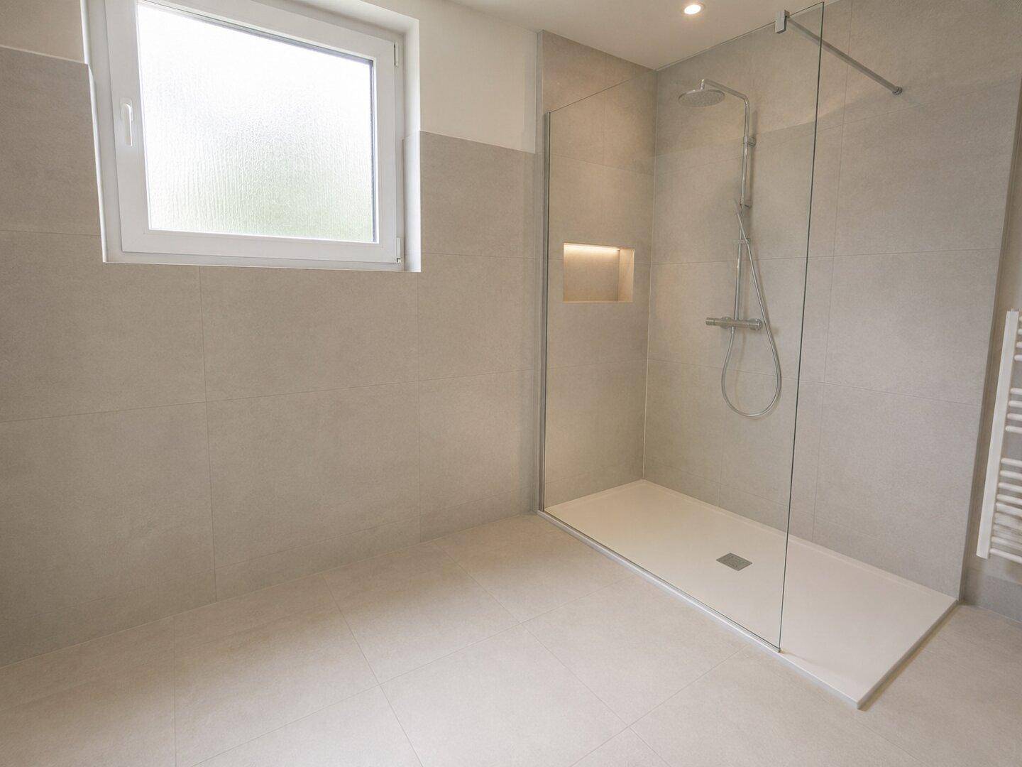 Modernes Badezimmer mit hellgrauen Fliesen, einer begehbaren Dusche und Fenster für Tageslicht.