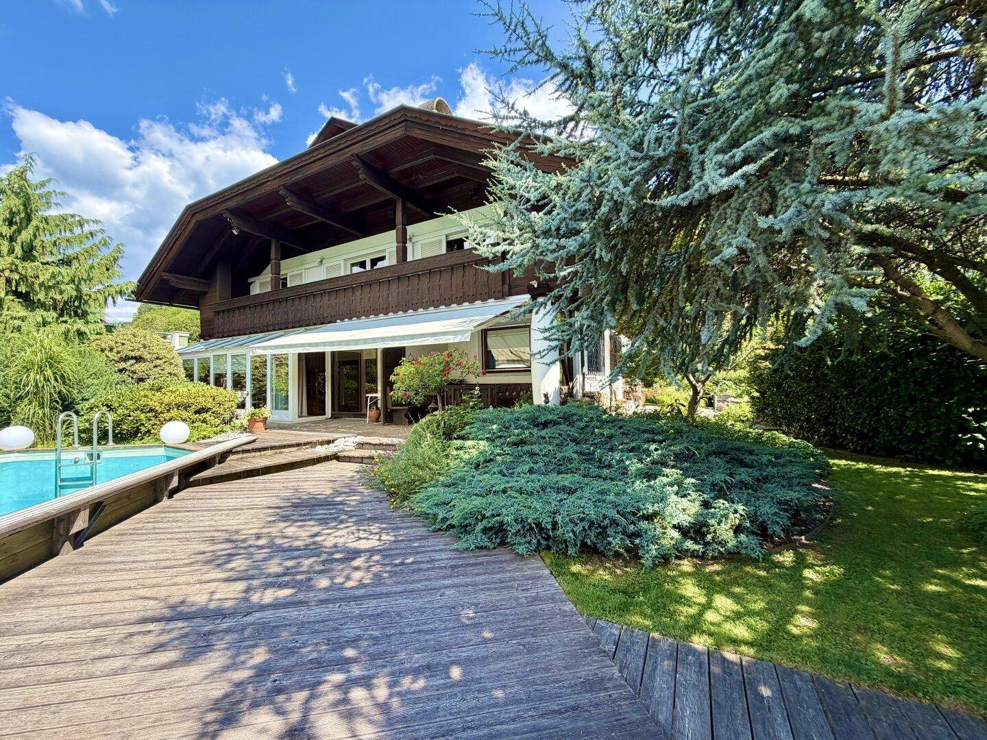 Hausfassade mit großem Holzdeck, Swimmingpool und üppiger Bepflanzung unter blauem Himmel.