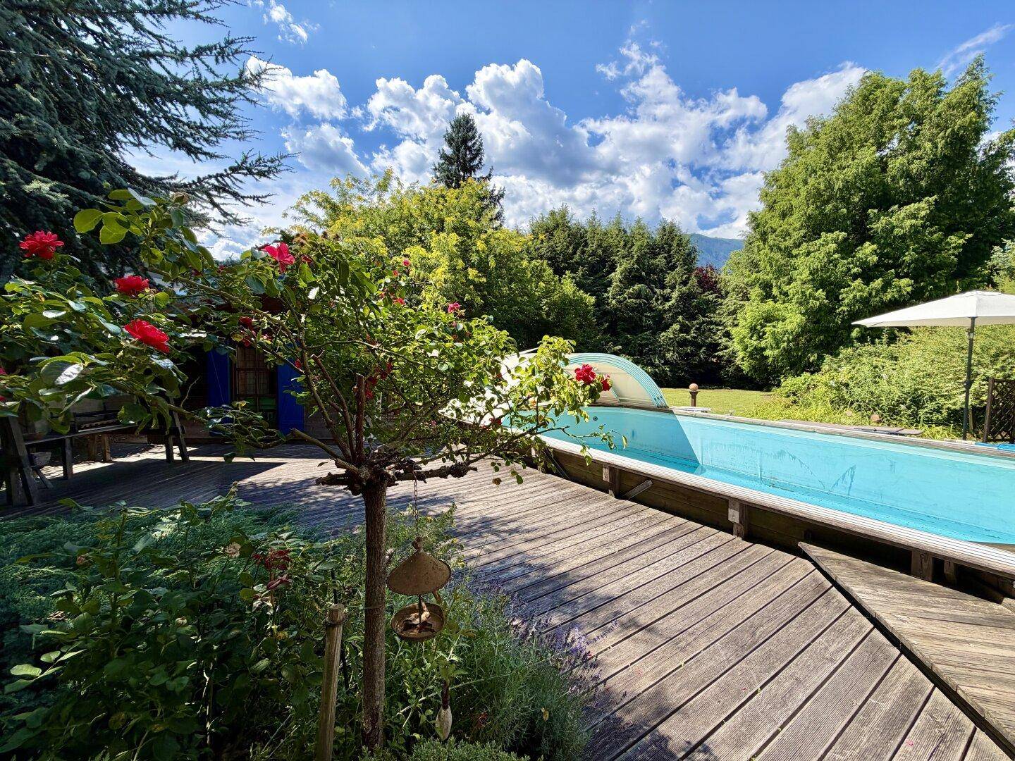 Gepflegter Gartenbereich mit Swimmingpool, Holzdeck und blühenden Rosen unter strahlend blauem Himmel.