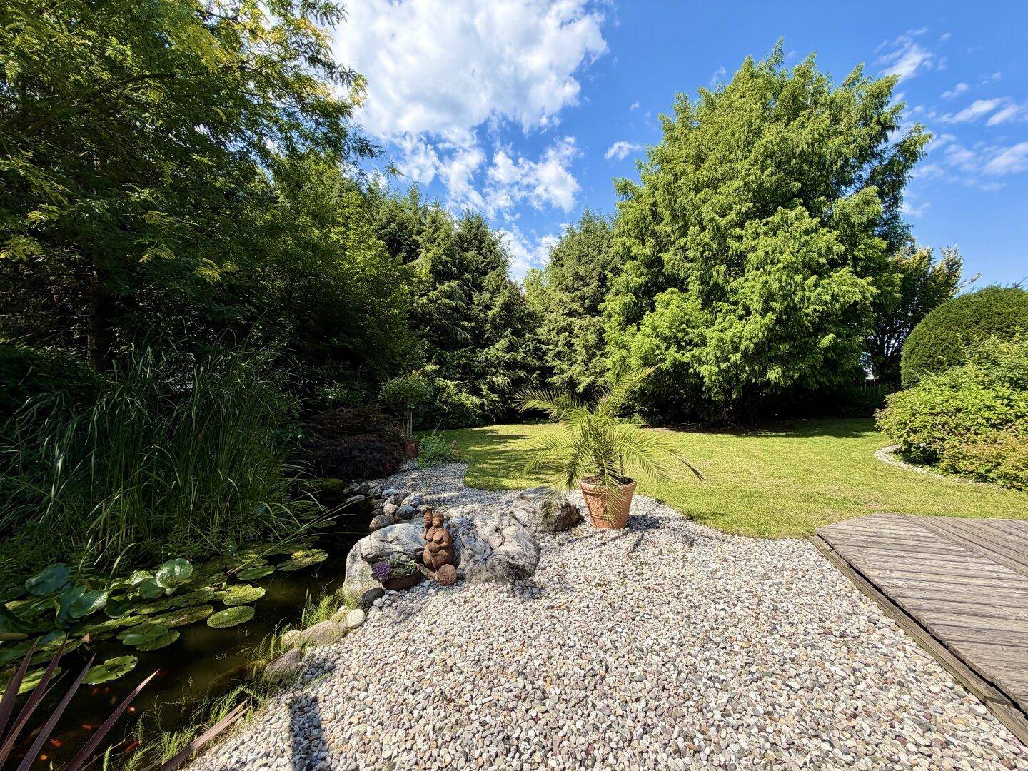 Idyllischer Gartenbereich mit Teich, Kiesbeet und üppiger grüner Vegetation unter blauem Himmel.