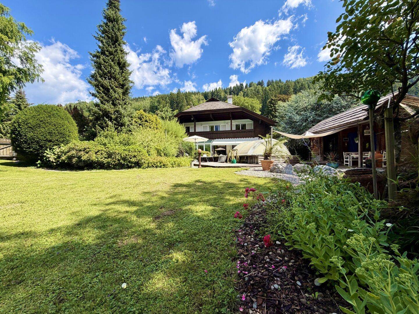 Weitläufiger Garten mit Rasenfläche und Blumenbeeten vor dem traditionellen Wohnhaus und bewaldeten Hügeln.