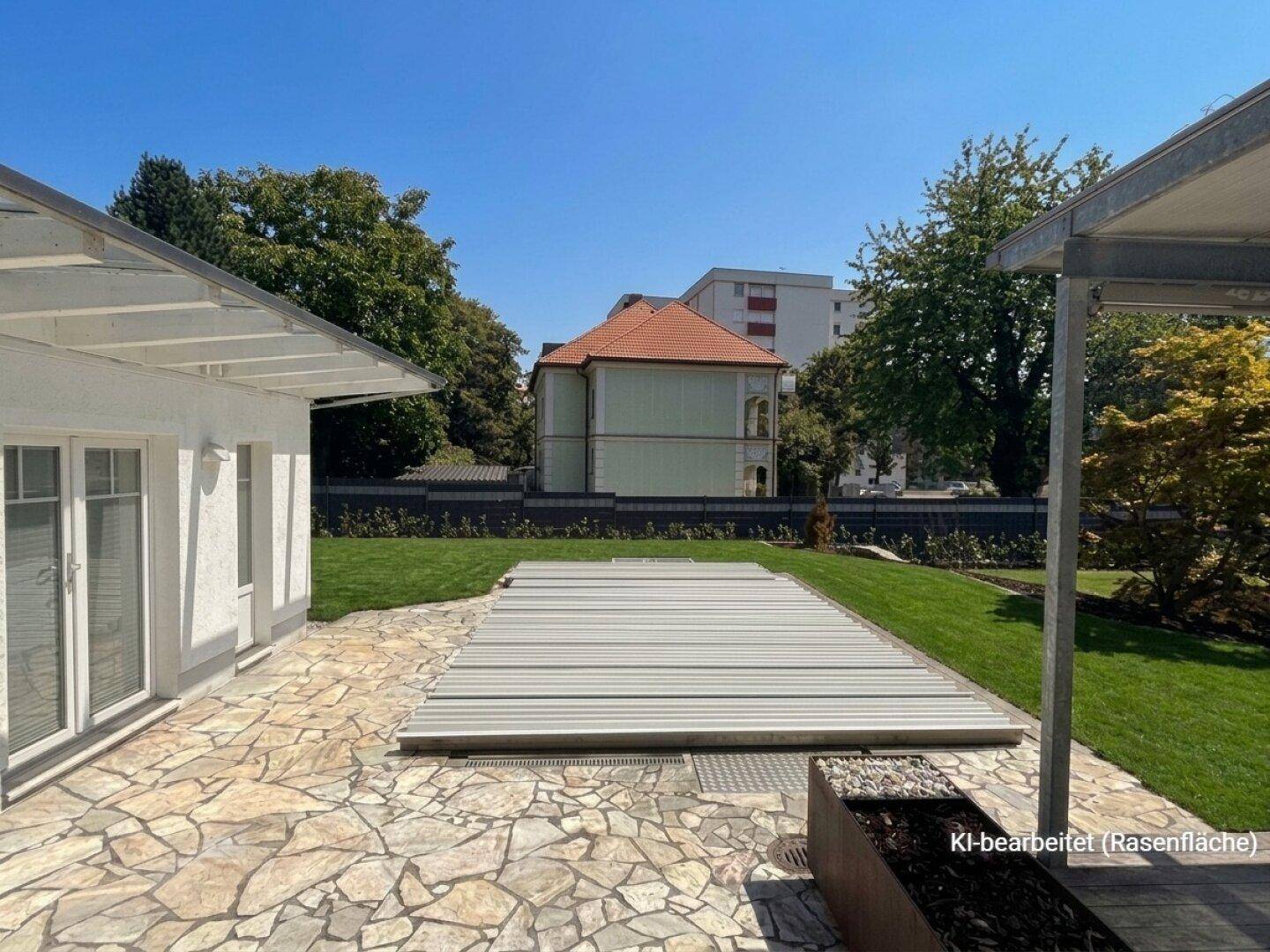 Gepflegter Garten mit Steinterrasse, großer Rasenfläche und einer Abdeckung, möglicherweise für einen Pool.