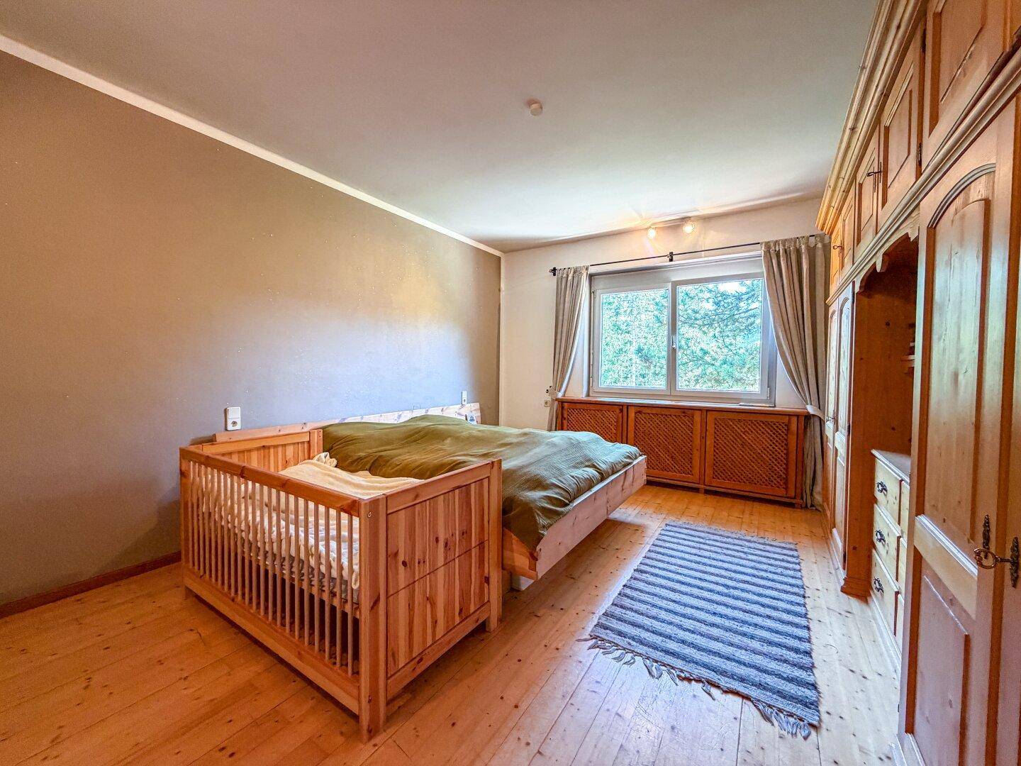 Helles Schlafzimmer mit Holzboden, großem Fenster und traditionellen Holzmöbeln, inklusive Babybett.