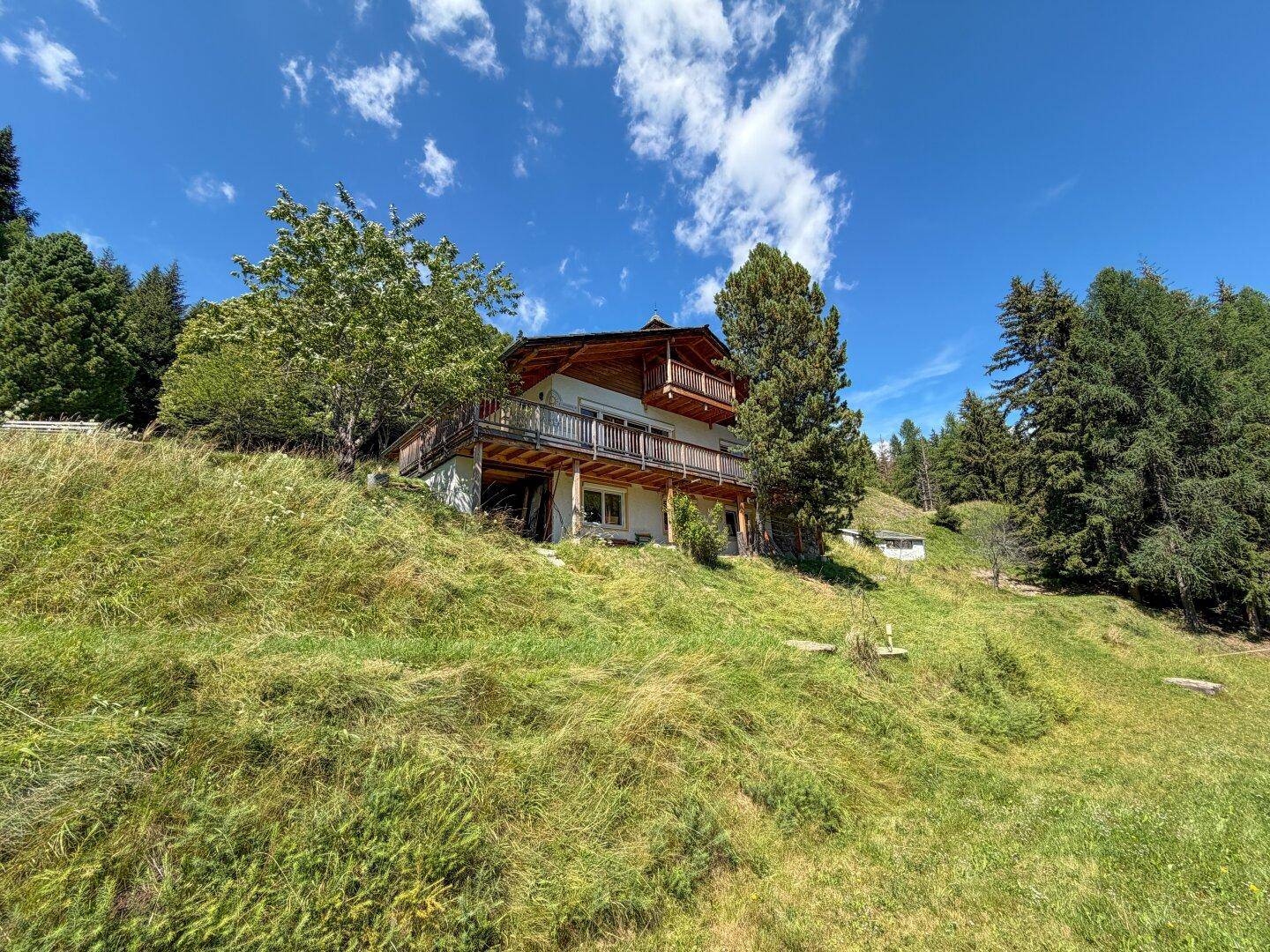 Alpines Holzhaus am Hang mit mehreren Balkonen, umgeben von grünen Wiesen und hohen Bäumen.