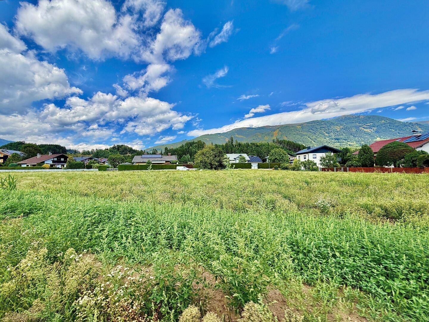 Weitläufiges grünes Feld mit Häusern und Bergen im Hintergrund unter blauem Himmel.