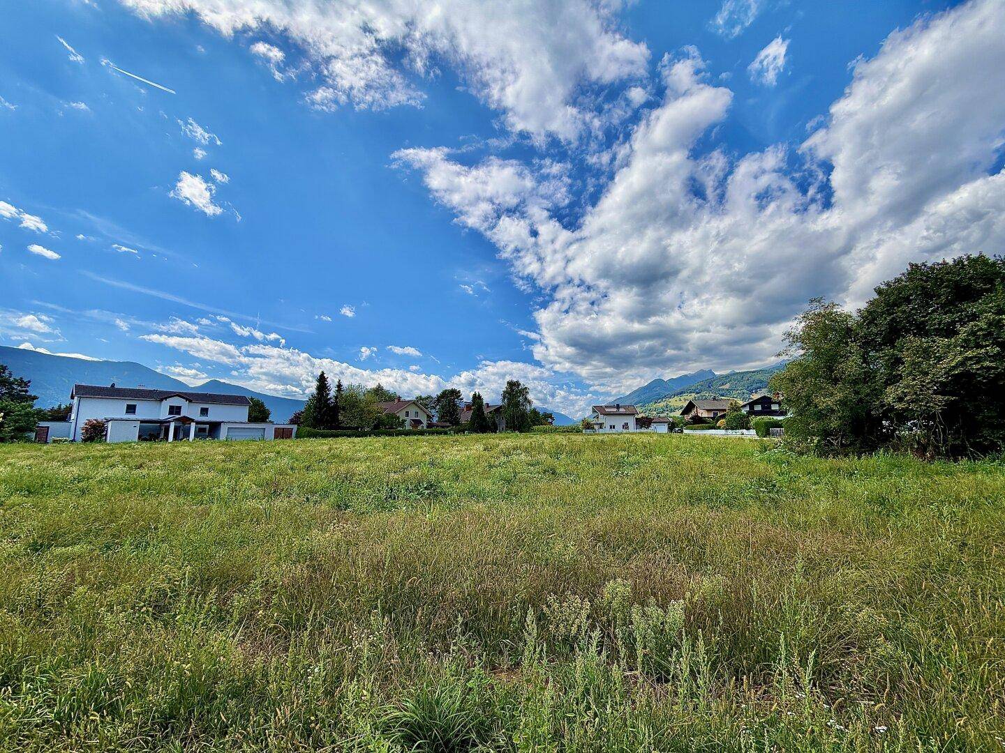 Grüne Wiese mit vereinzelten Häusern und einer Bergkette am Horizont unter blauem Himmel.