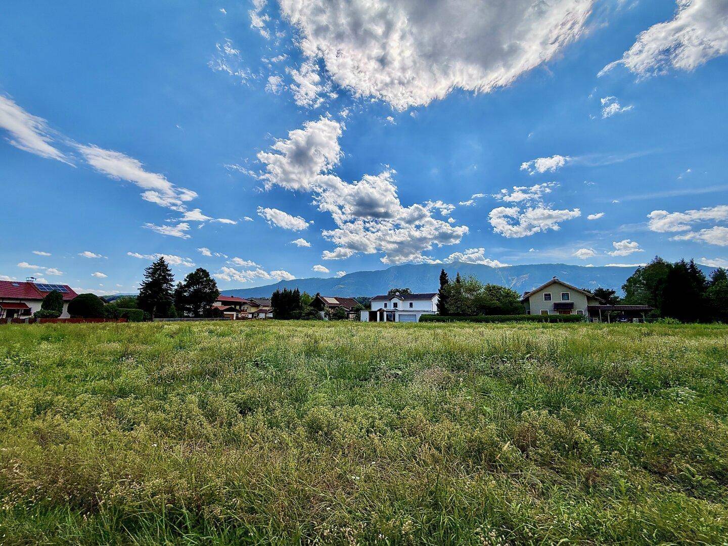 Weitläufiges grünes Feld mit Häusern und Bergen in der Ferne unter einem strahlend blauen Himmel.