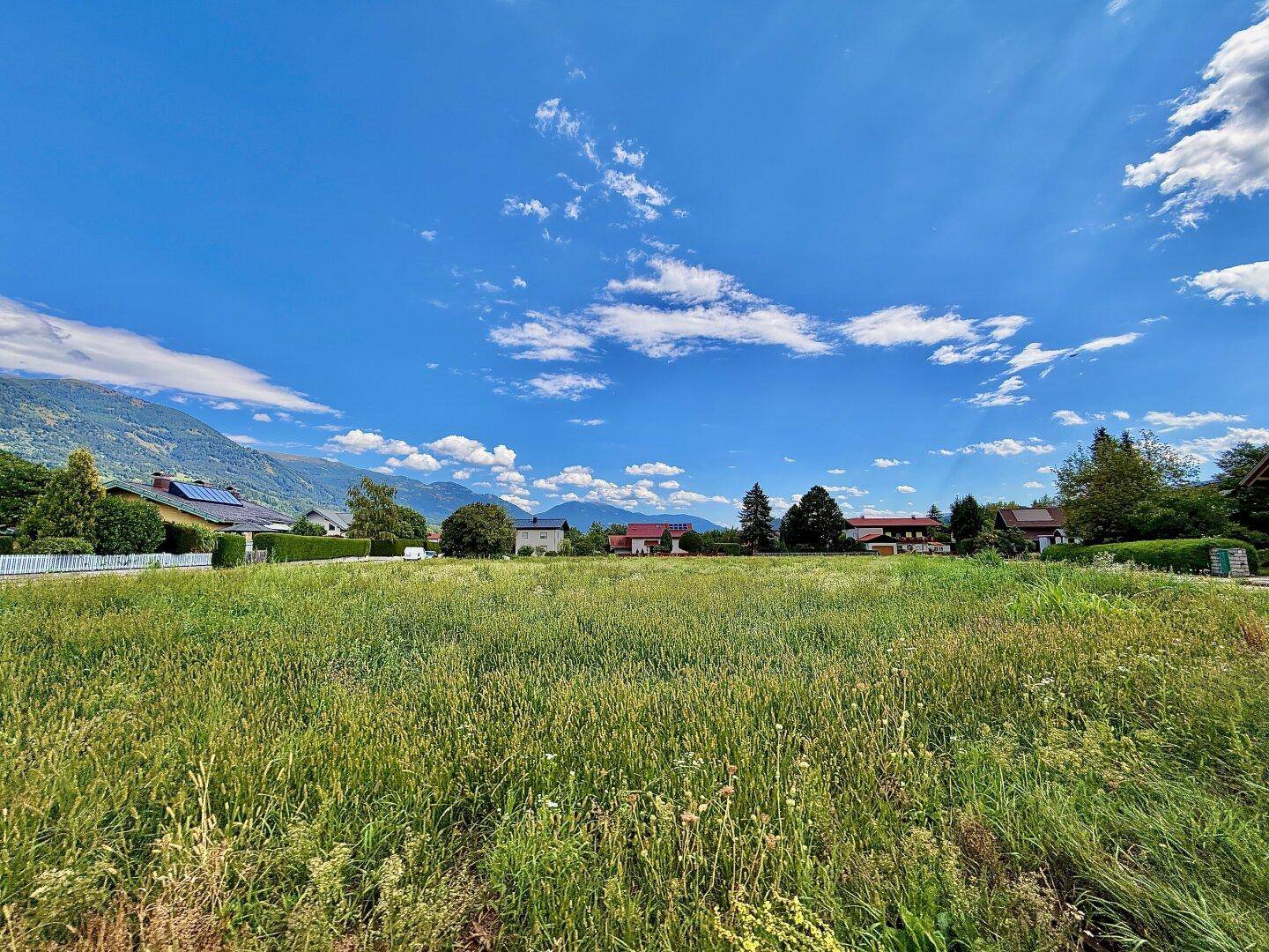 Großes grünes Feld mit Blick auf Häuser und die umliegende Berglandschaft bei Sonnenschein.
