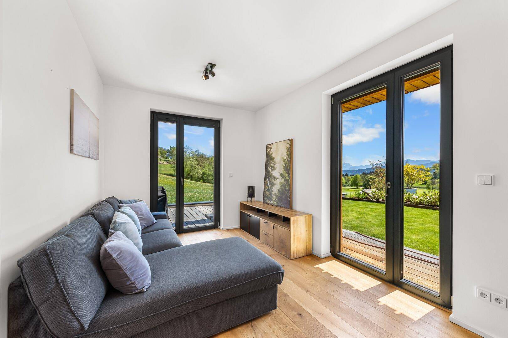 Gemütliches Wohnzimmer mit Sofa, Holzboden und großen Fenstern mit Blick auf den Rasen.