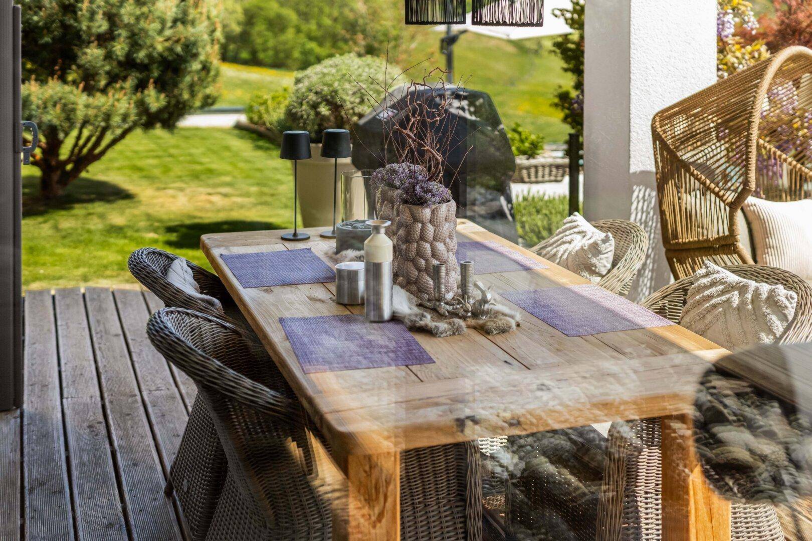 Detailaufnahme eines großen Holztisches mit Korbstühlen auf einer sonnigen Terrasse.