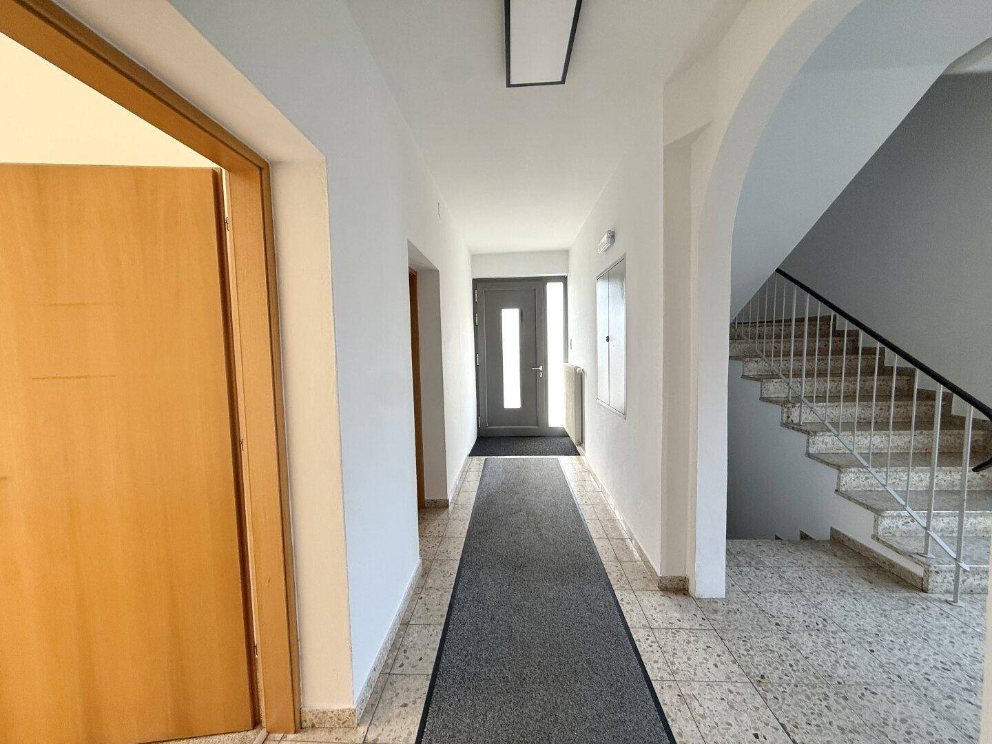 Heller Flur mit gefliestem Boden, Treppenaufgang und einer modernen Eingangstür am Ende.