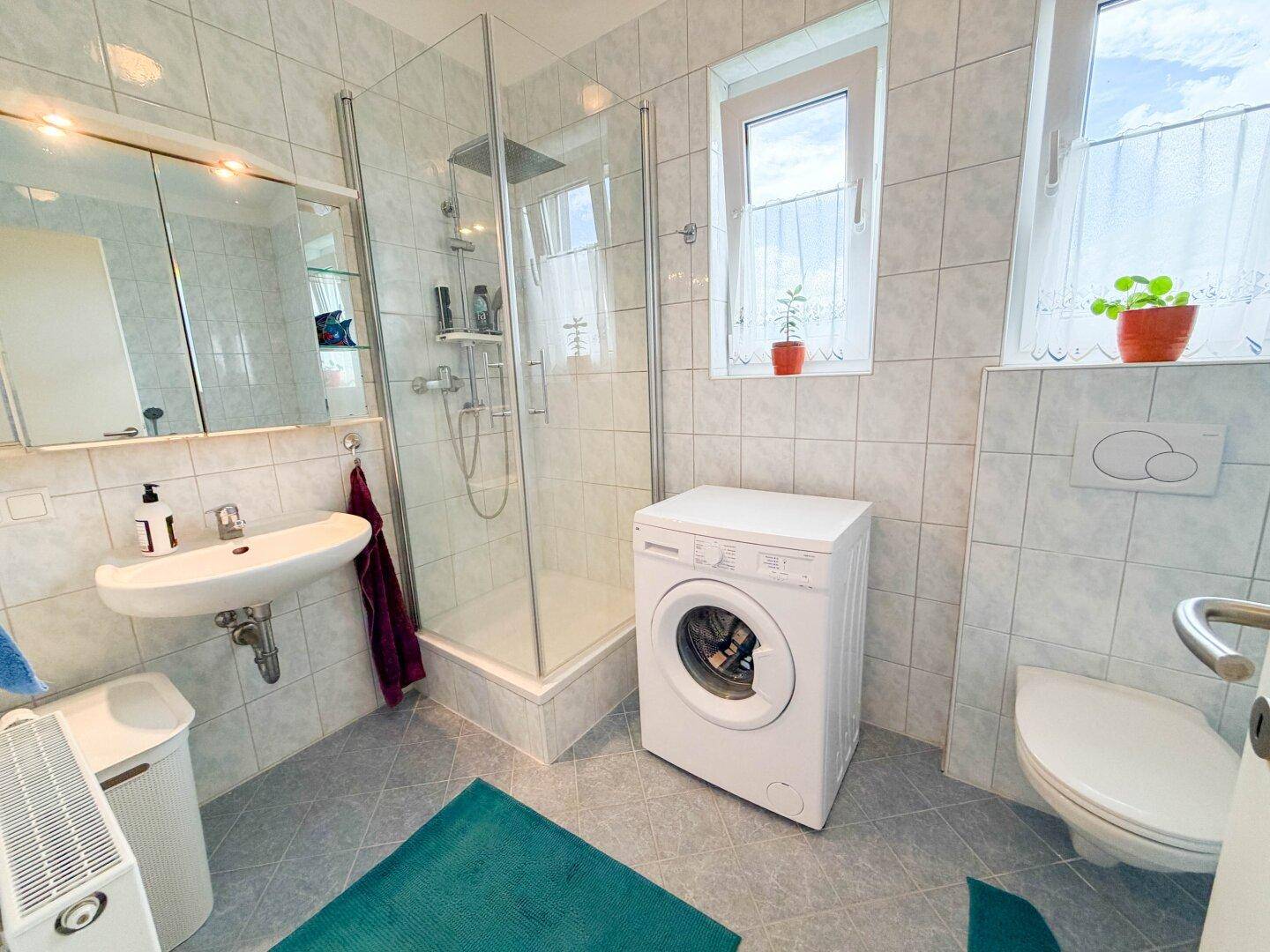Helles Badezimmer mit Dusche, Waschbecken, WC und Waschmaschine, gefliest.