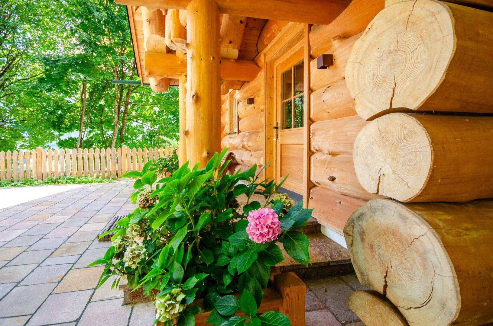 Eingangsbereich des Holzhauses mit gepflasterter Terrasse und blühenden Hortensien, umgeben von einem Holzzaun.