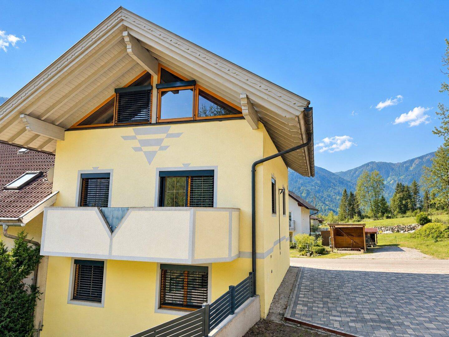 Seitenansicht des gelben Hauses mit Balkon, gepflasterter Fläche und Bergblick.