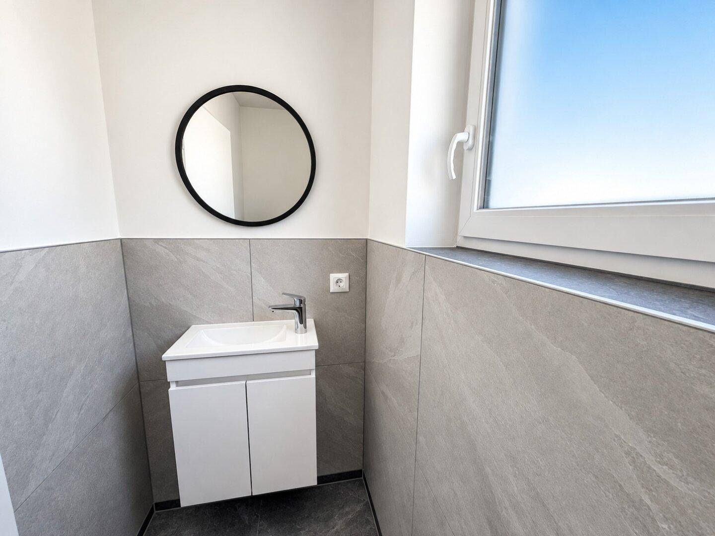 Kleines Gäste-WC mit modernem Waschbecken, rundem Spiegel und Fenster.