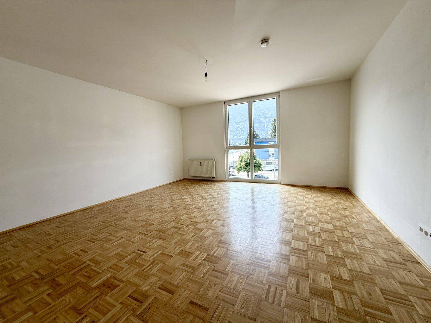 Helles Wohnzimmer mit Fischgrätparkettboden und großem Fenster mit Blick ins Grüne.