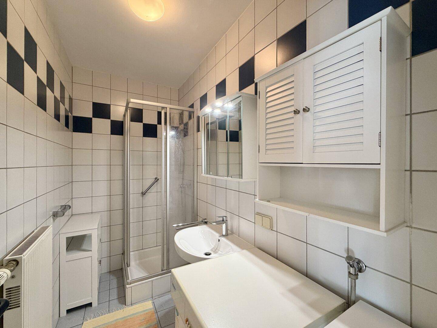 Gefliestes Badezimmer mit Duschkabine, Waschbecken, Spiegelschrank und Waschmaschine.