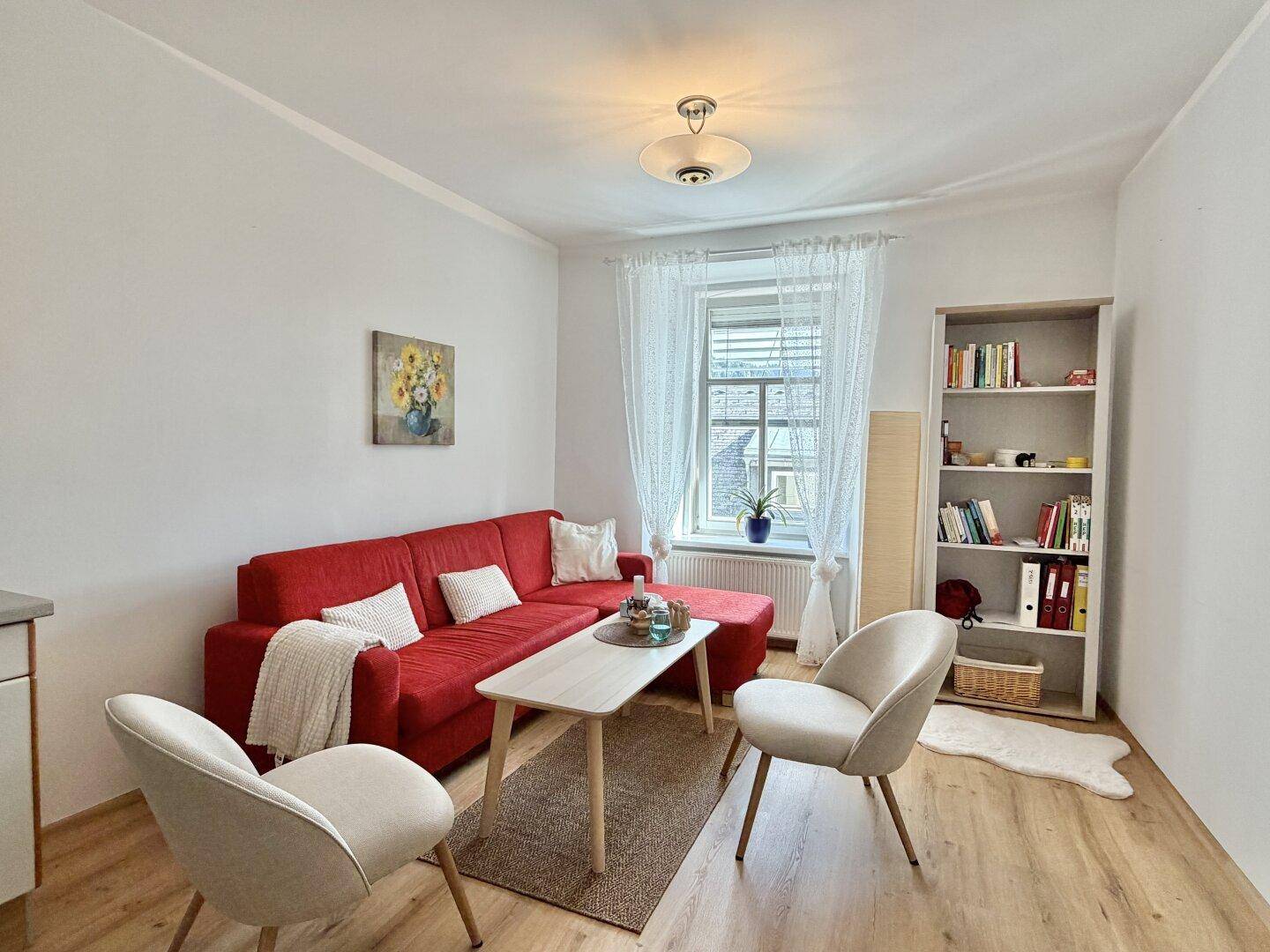 Gemütliches Wohnzimmer mit rotem Ecksofa, zwei Sesseln und einem Bücherregal am Fenster.