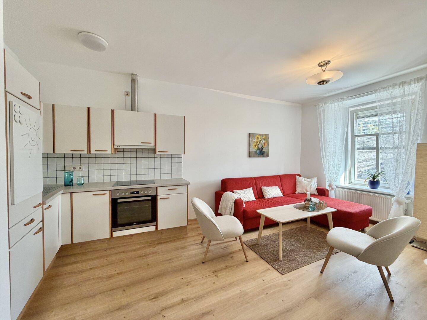 Offener Wohnbereich mit integrierter Küche, rotem Sofa und hellen Küchenschränken.