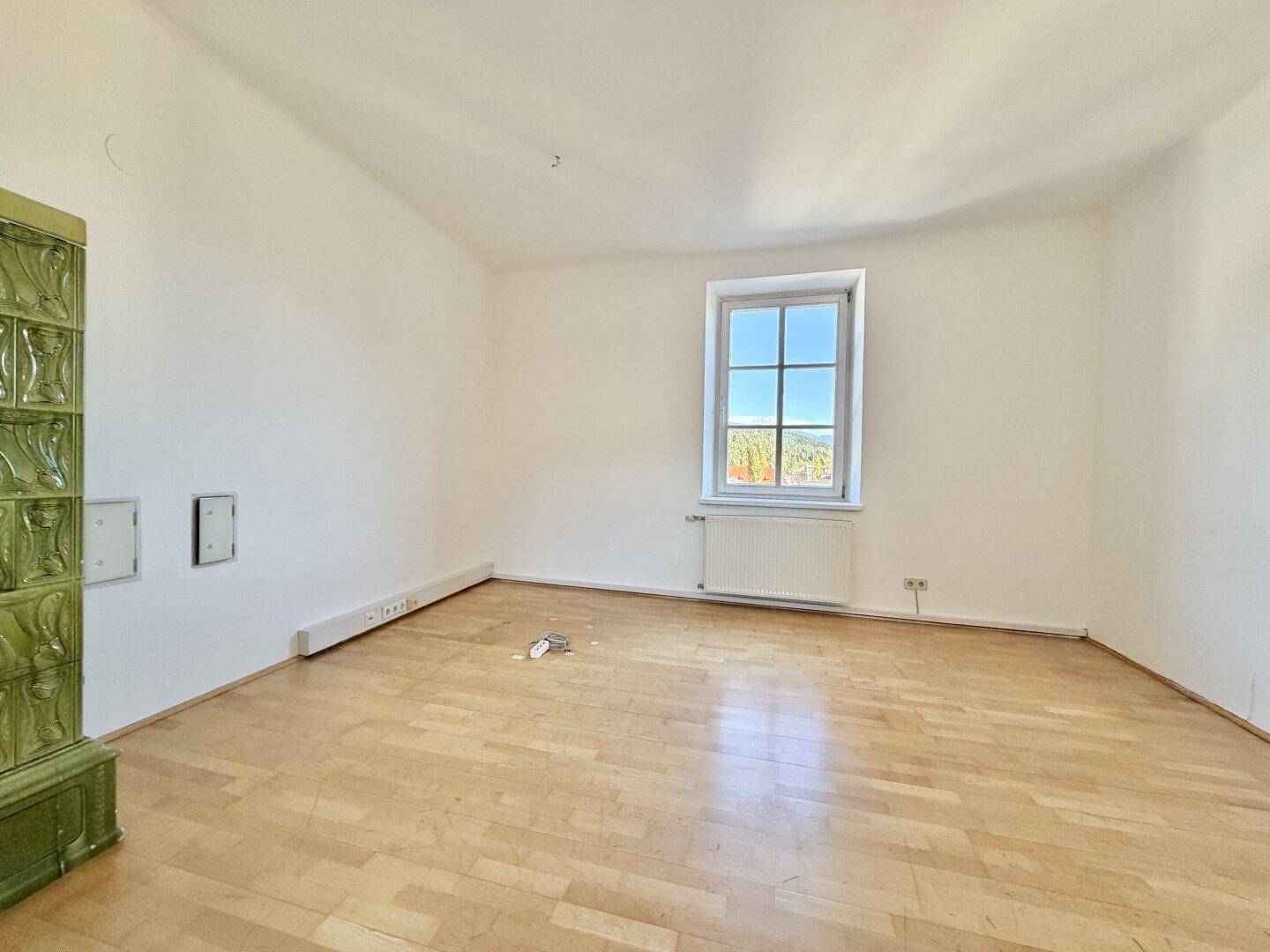 Heller Raum mit Laminatboden, einem grünen Kachelofen und einem Fenster mit Blick ins Grüne.
