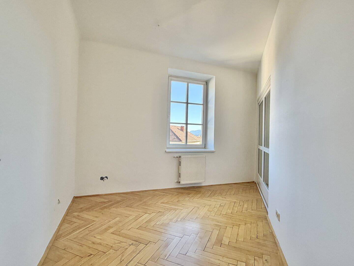 Gemütlicher Raum mit Fischgrätparkett, weiß gestrichenen Wänden und einem Fenster mit Ausblick.