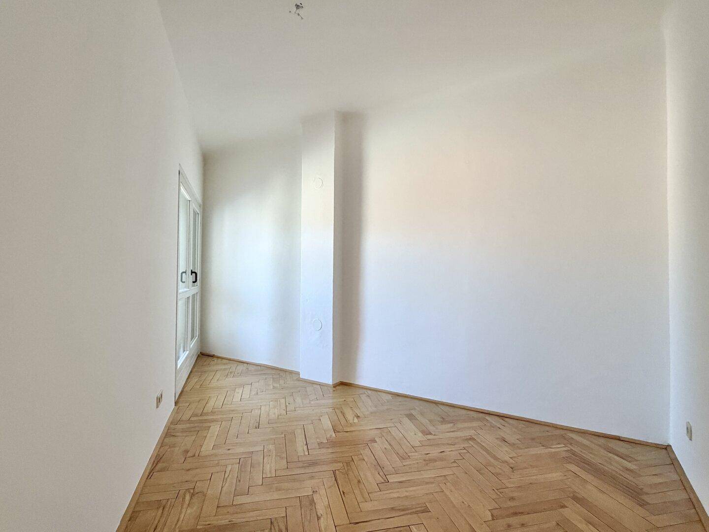 Heller Raum mit Fischgrätparkettboden und einer Glastür, die zu einem weiteren Bereich führt.