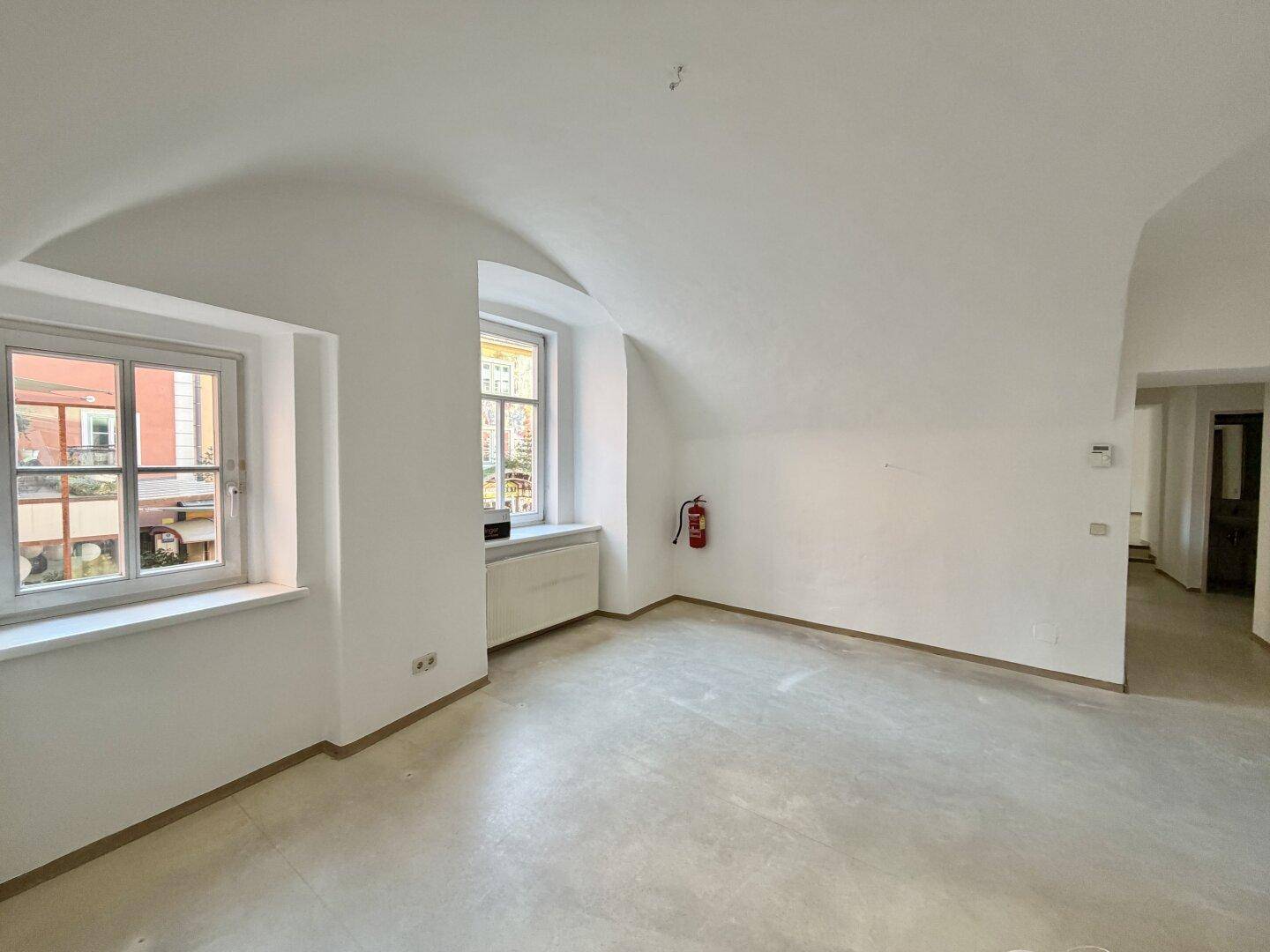 Heller Raum mit Gewölbedecke, zwei Fenstern und einem Feuerlöscher an der Wand.