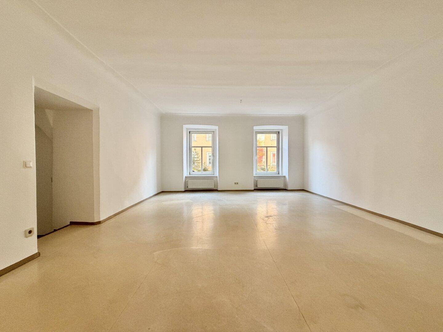 Großer, heller Raum mit hellem Holzboden und zwei Fenstern, die viel Licht spenden.