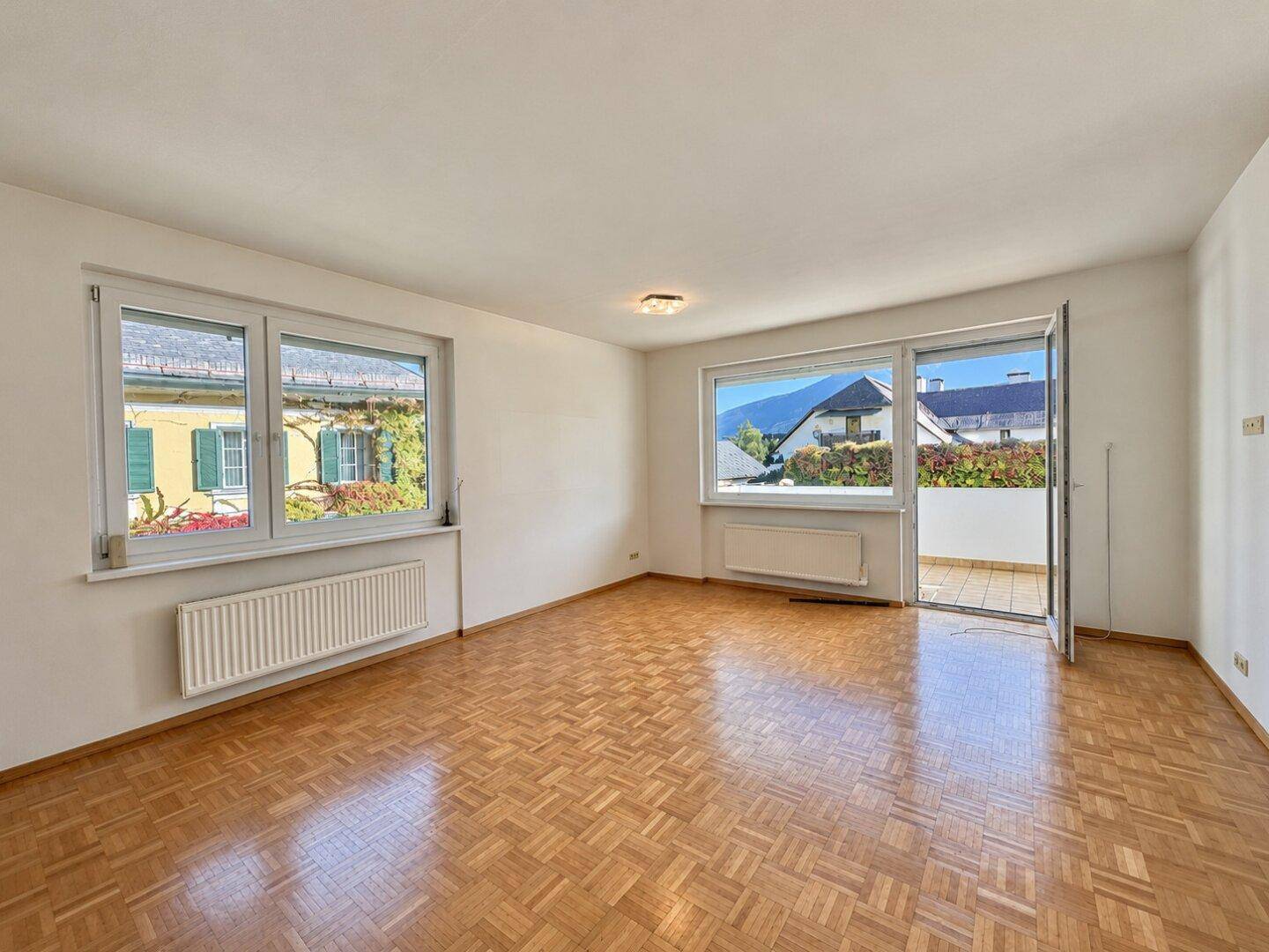 Helles Wohnzimmer mit Parkettboden, großen Fenstern und Zugang zu einem Balkon.