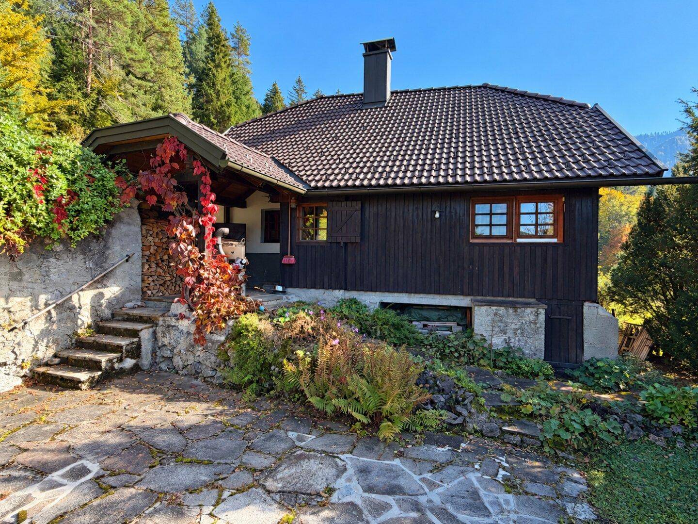 Rustikales Haus mit dunkler Holzfassade, Steintreppe und herbstlicher Bepflanzung am Eingang.