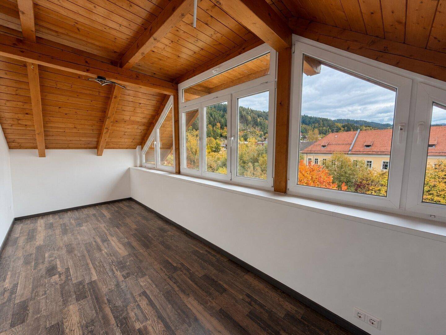 Heller Dachbodenraum mit Holzbalkendecke, großen Fenstern und Blick auf die herbstliche Landschaft.