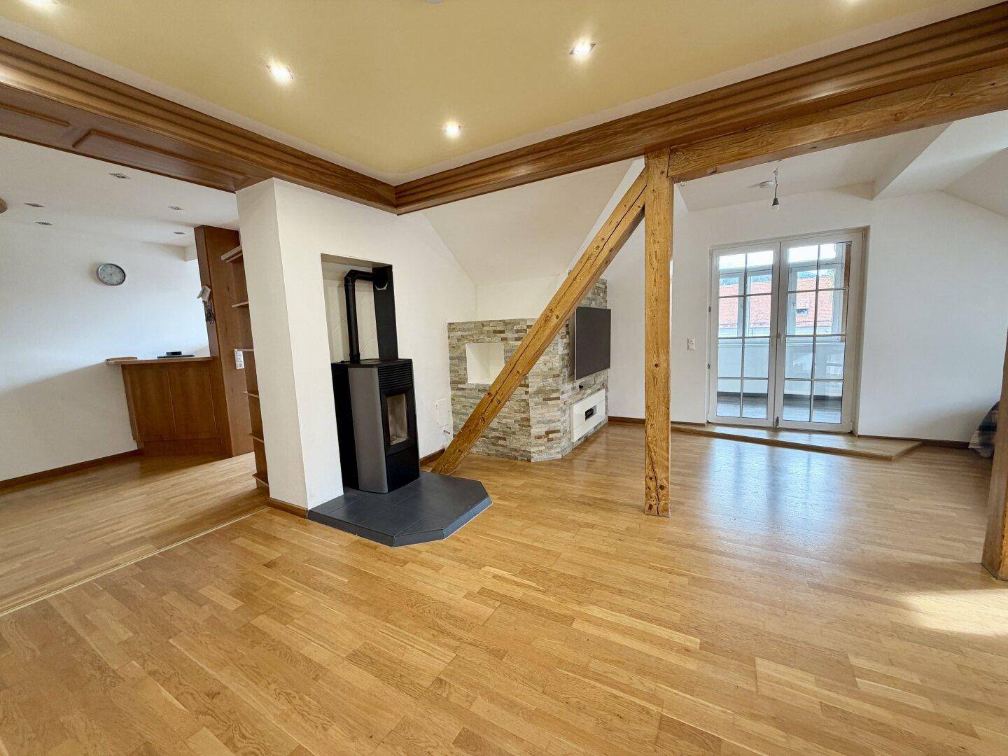 Heller Wohnbereich mit Holzbalken, Parkettboden, modernem Kaminofen und Zugang zu einem Balkon.