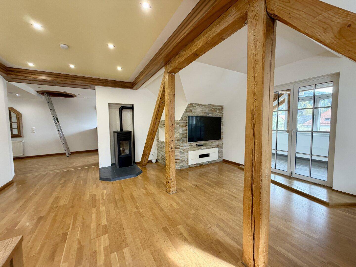 Geräumiges Wohnzimmer mit Holzbalken, Parkettboden, modernem Kaminofen und Zugang zu einem Balkon.